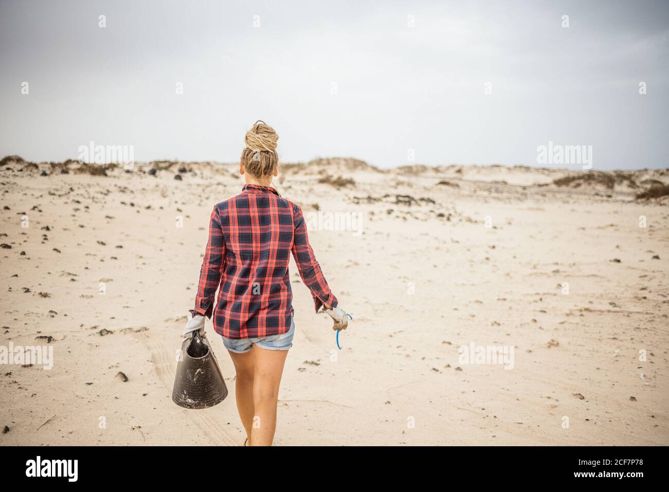 Positive hipster femme dans des vêtements décontractés et des gants de collecte de déchets dans le sac tout en s'accroupidant sur la plage déserte Banque D'Images