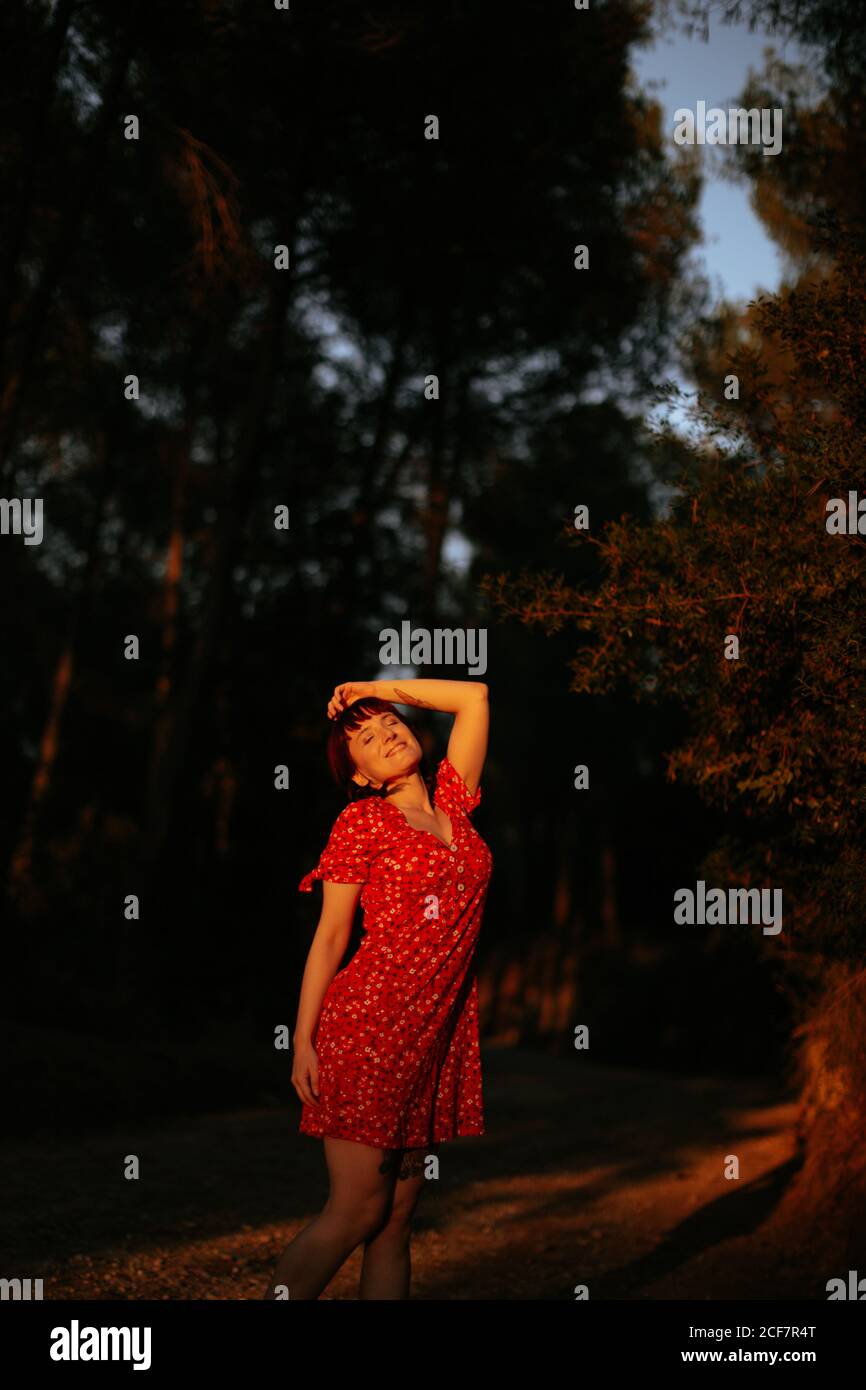 Femme gaie et attentionnés vêtue de robe rouge, souriante au milieu de grands arbres le jour ensoleillé dans la forêt verte avec les yeux fermés Banque D'Images