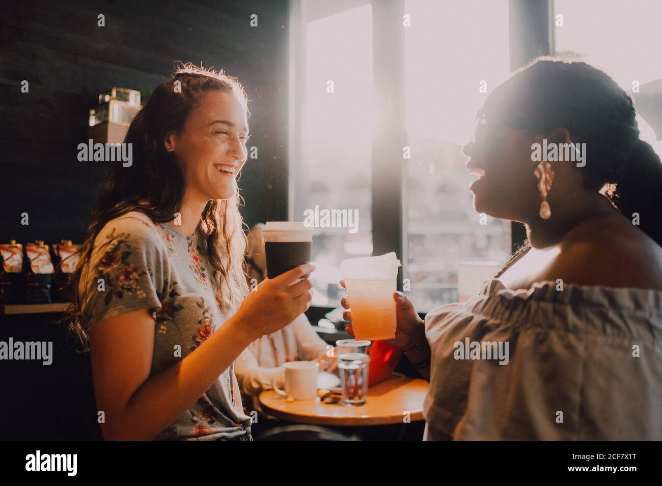 Vue latérale de gaie multiraciale jeune décontracté femmes riant et boire du café tout en étant assis près de la fenêtre au café au coucher du soleil Banque D'Images
