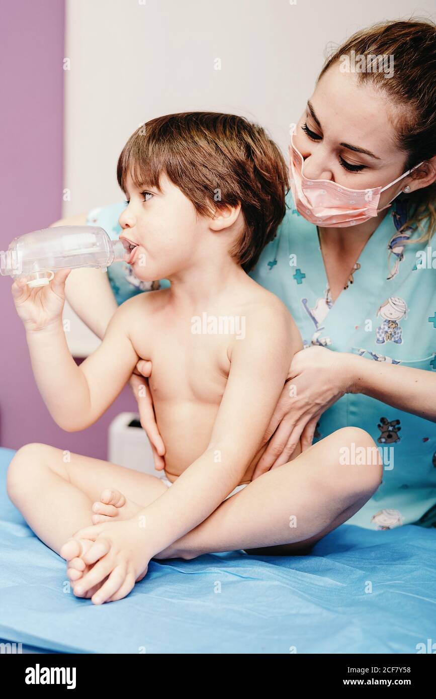 Un petit garçon atteint d'une maladie respiratoire utilisant un inhalateur alors qu'il est jeune et amical une infirmière féminine se tenant à proximité et apportant son soutien à la clinique Banque D'Images