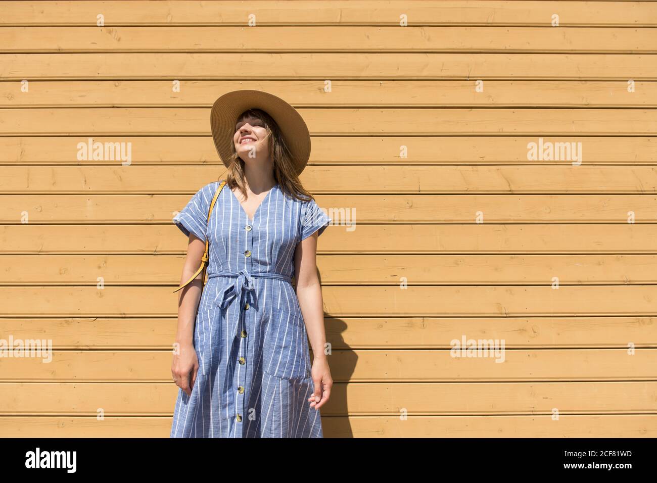 Jolie femme souriante dans un chapeau de paille et une sundress debout avec les yeux fermés devant un mur en bois Banque D'Images