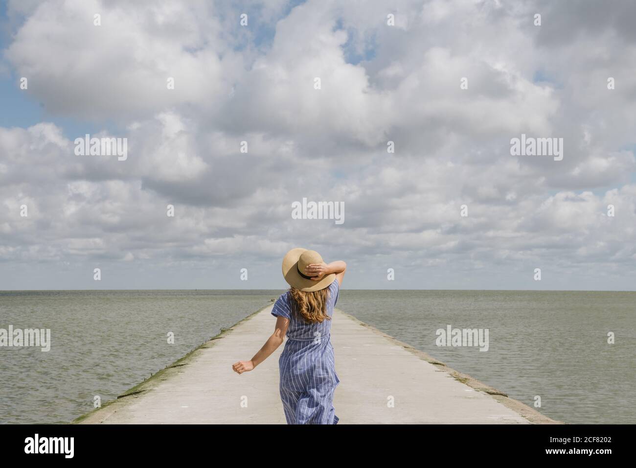 Vue arrière d'une femme adulte en chapeau de paille et de la sundress qui court sur un quai en béton vide par temps nuageux Banque D'Images