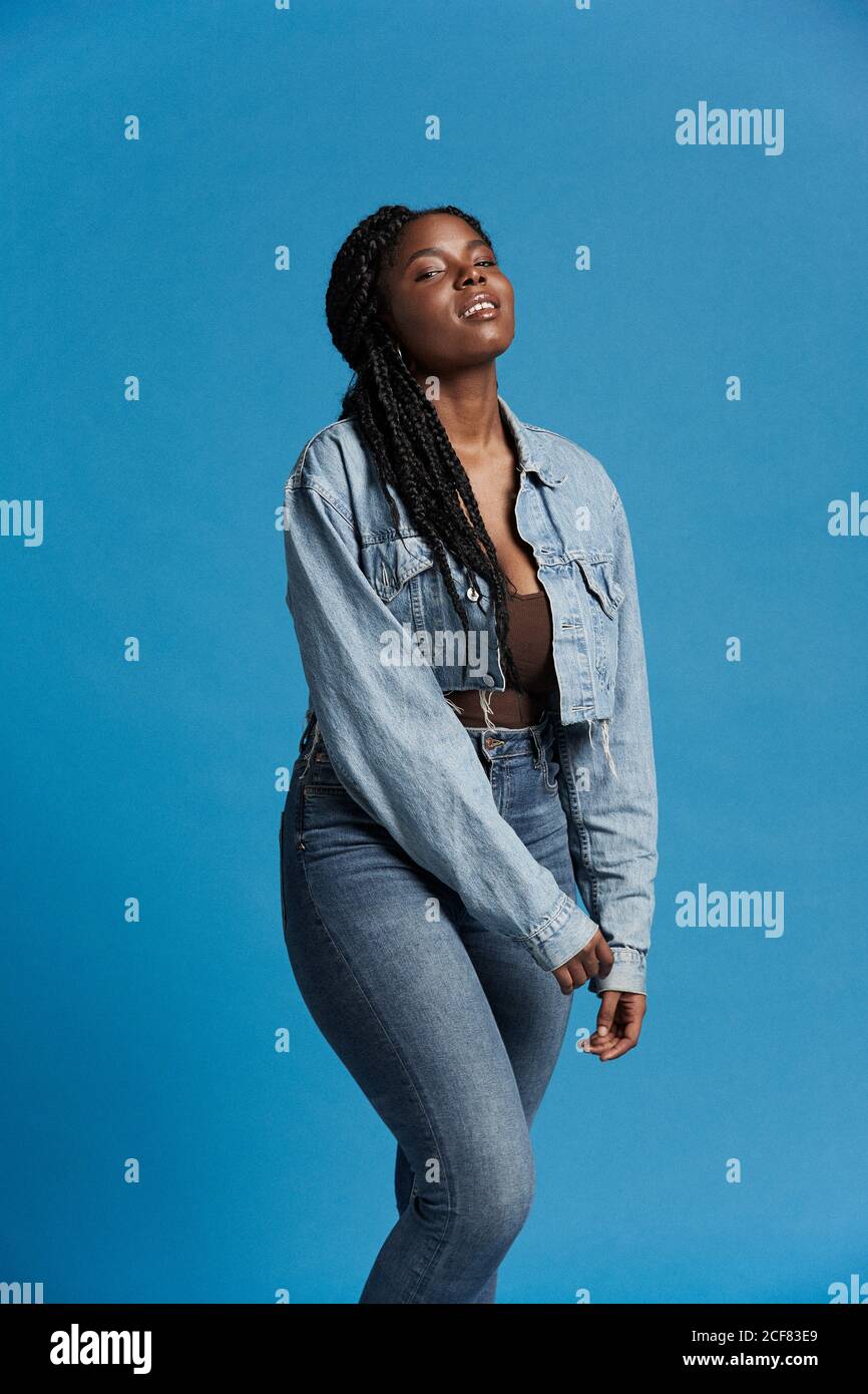 Positive belle Afro-américaine femme avec des tresses dans une tenue en denim souriant et regardant l'appareil photo sur fond bleu Banque D'Images