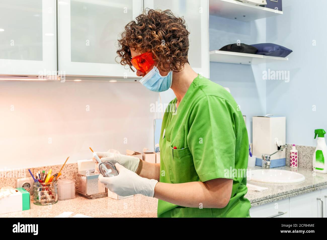 Vue latérale d'un spécialiste spécialisé concentré en uniforme vert et lunettes de protection rouges pour la préparation de l'outil dentaire pour le remplissage des dents Banque D'Images