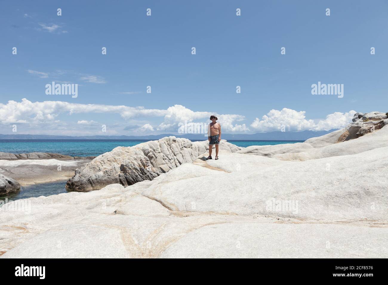 Homme mature bronzé debout sur un rivage rocheux blanc avec ciel bleu et nuages, Halkidiki, Grèce Banque D'Images