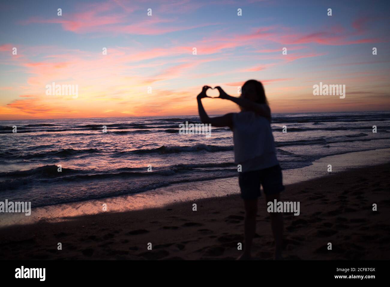 Femme faisant un geste de coeur pendant la marche sur la plage de sable contre ciel pittoresque au coucher du soleil Banque D'Images