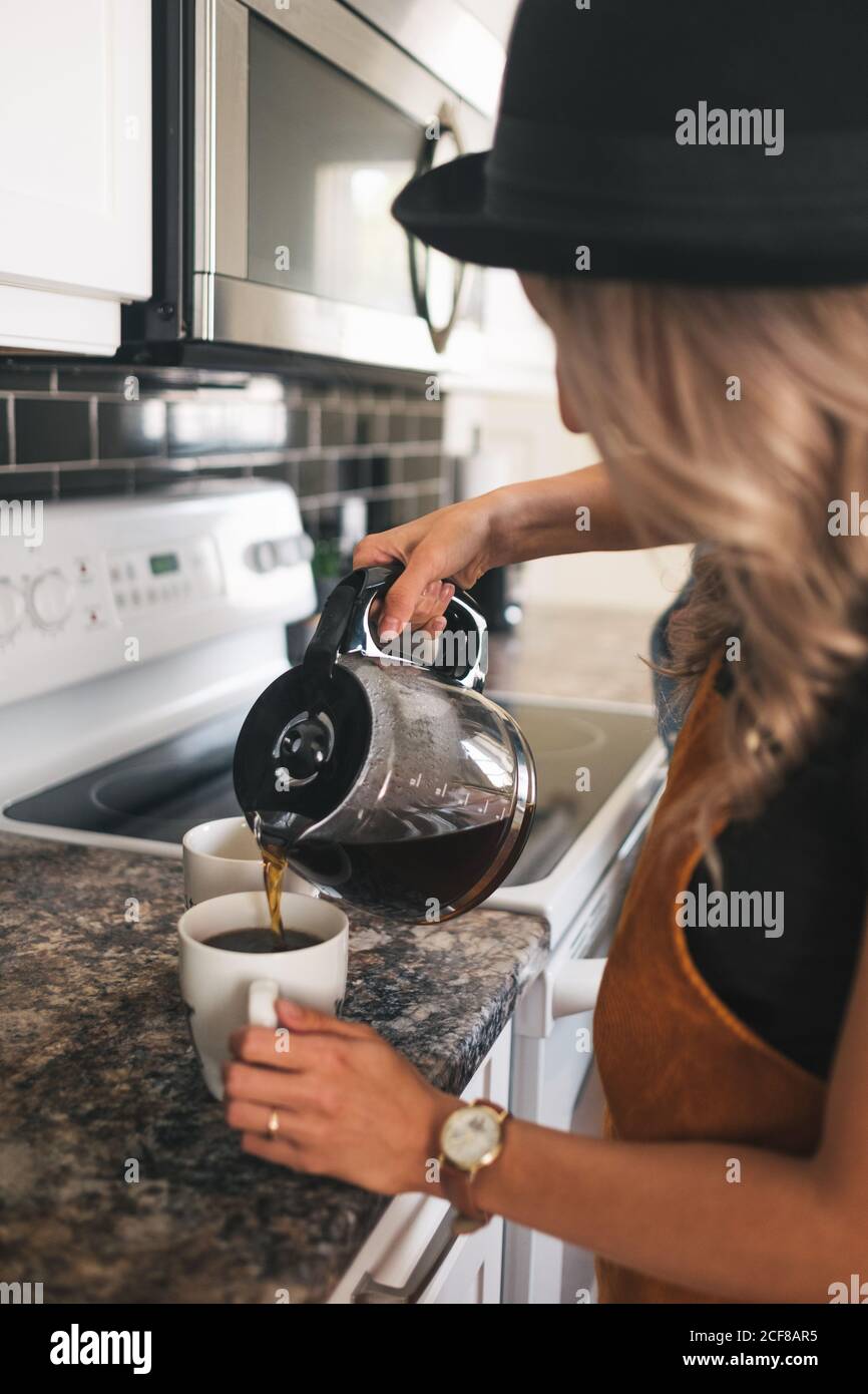 Vue latérale rognée d'une femme méconnue dans un chapeau avec montre-bracelet versez du café fraîchement préparé dans des tasses blanches en vous tenant debout cuisine à la maison Banque D'Images