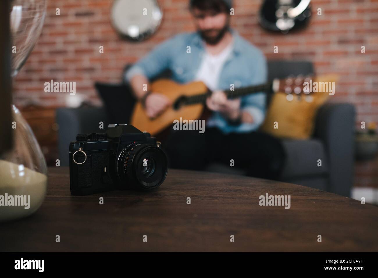 Crop Bearded gars dans la veste de denim jouant de la guitare acoustique pendant asseyez-vous sur un canapé confortable devant une table en bois avec appareil photo et sablier à la maison Banque D'Images