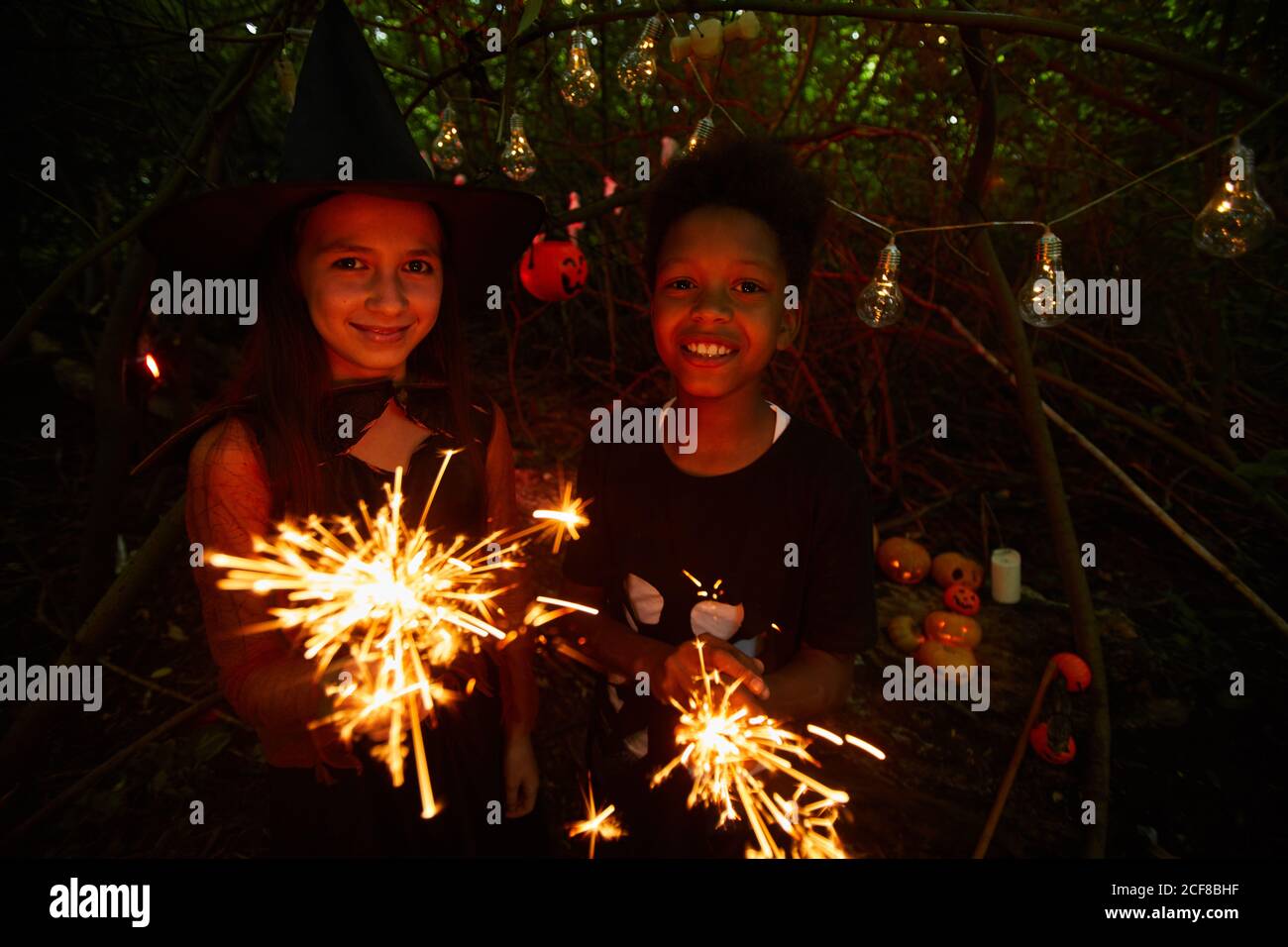 Portrait d'amis tenant des étincelles et souriant à l'appareil-photo pendant debout dans la forêt sombre et célébrant Halloween ensemble Banque D'Images