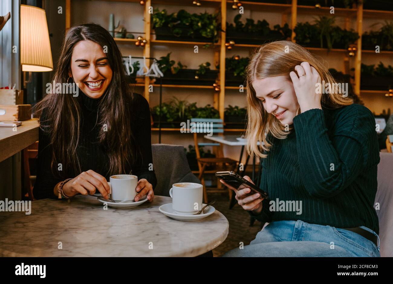 Les jeunes femmes rient à la blague tout en buvant du café et en utilisant smartphone pendant la réunion dans un restaurant confortable Banque D'Images