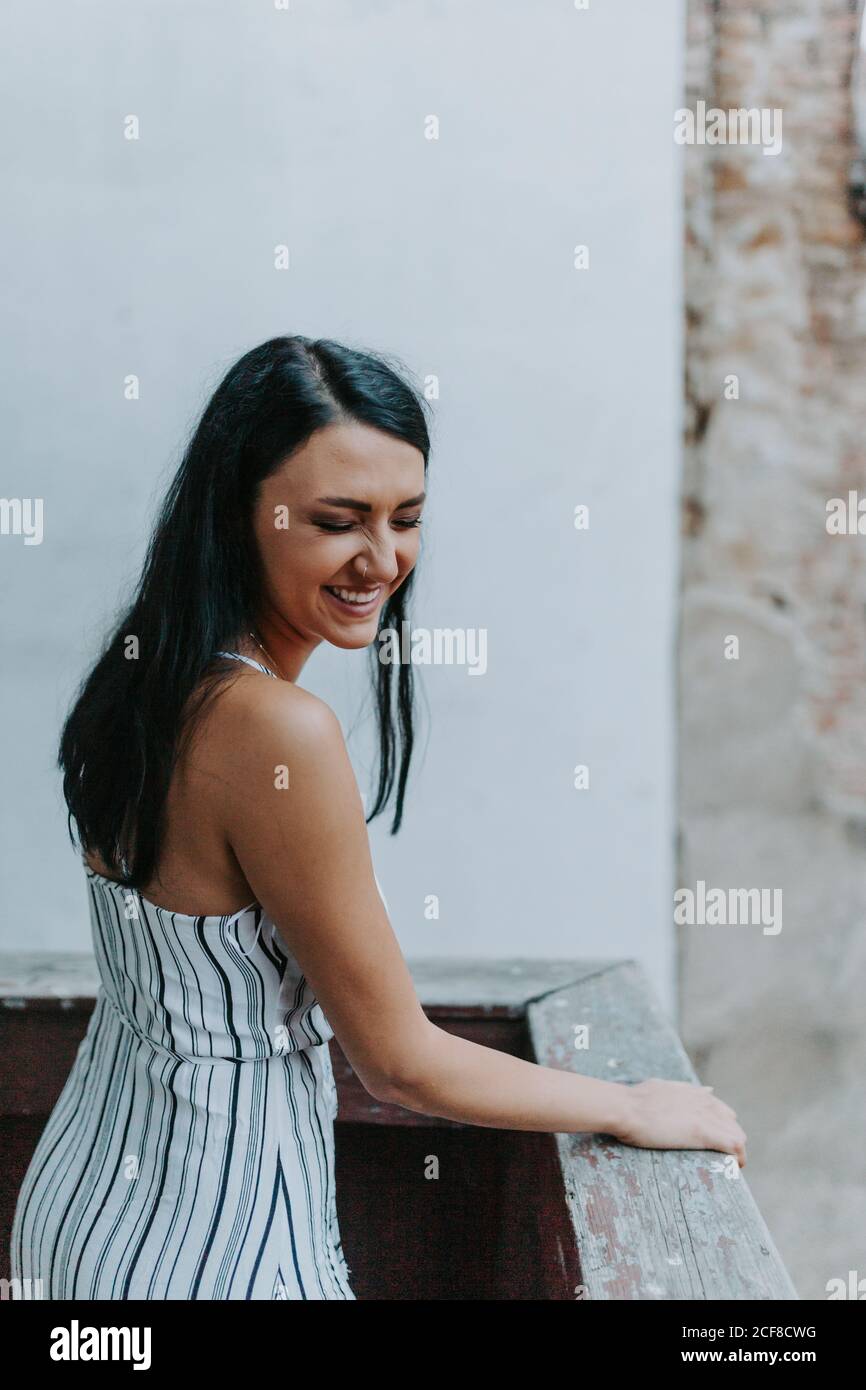 Jeune femme en robe rayée qui regarde loin en étant debout balcon d'un bâtiment vieux et miteux Banque D'Images