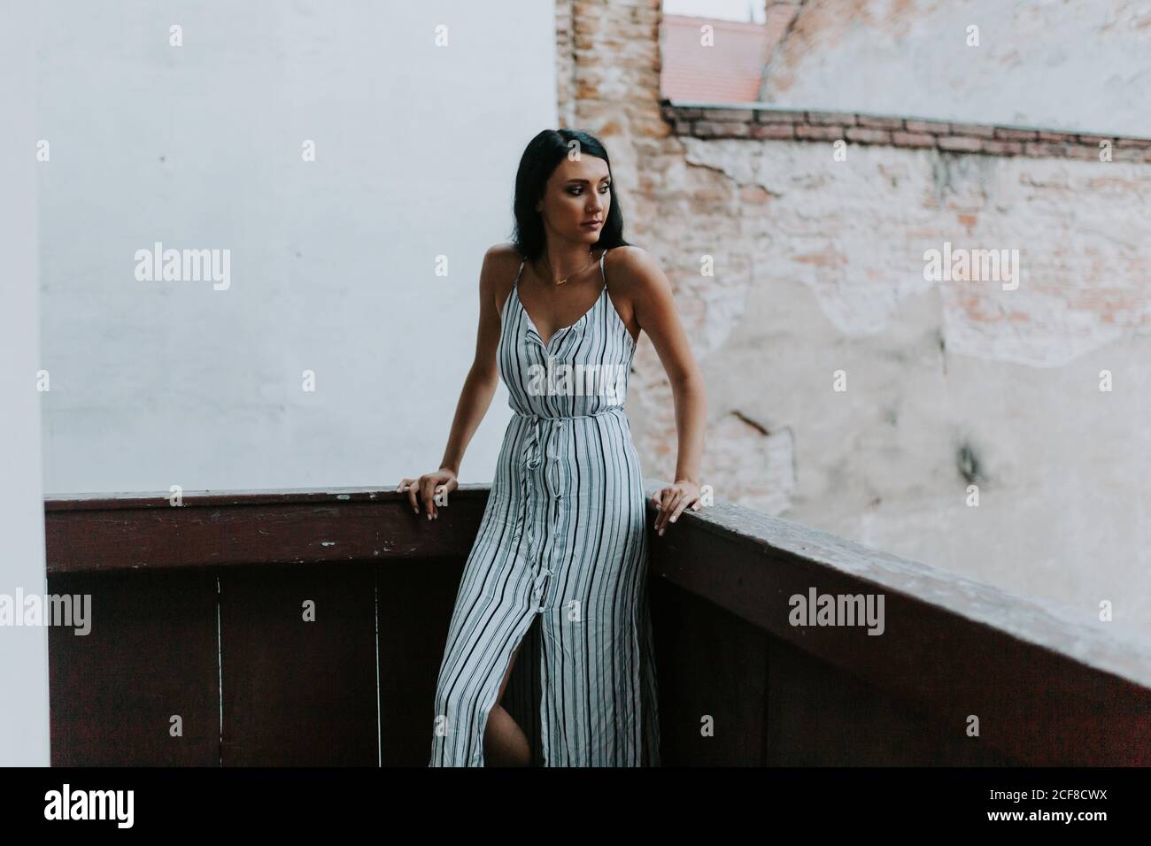 Jeune femme en robe rayée qui regarde loin en étant debout balcon d'un bâtiment vieux et miteux Banque D'Images