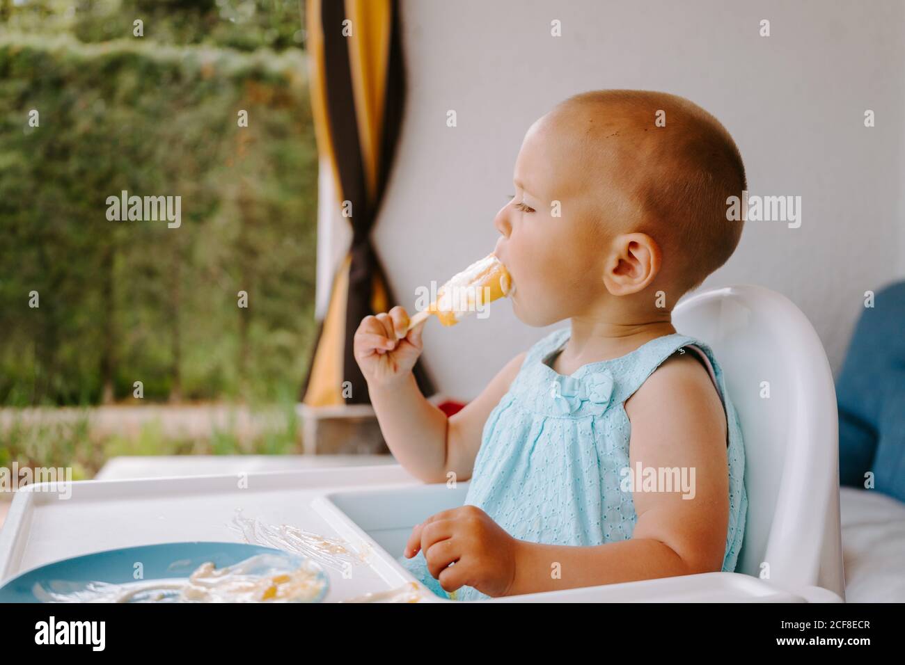 Un tout-petit mignon assis sur une chaise haute sur la terrasse et en train de manger de délicieuses glaces tout en regardant loin Banque D'Images