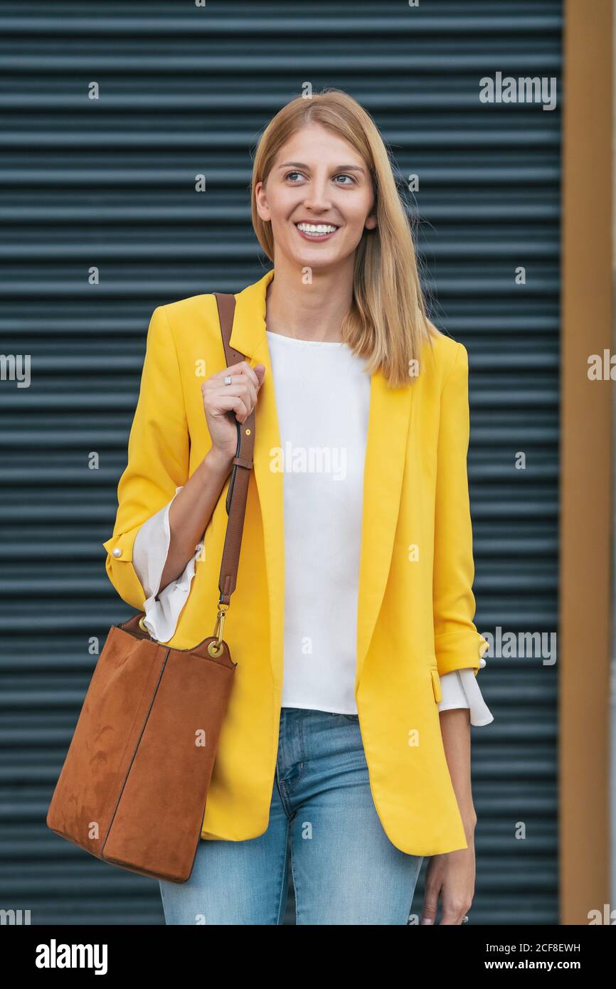 Bonne jeune femme blonde en blouson et Jean jaune vif avec un sac à main marron sur l'épaule en souriant tout en restant debout rue contre un mur à rayures floues en ville Banque D'Images