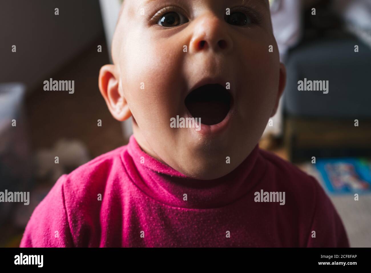 Petit garçon stupéfié petit dans une chemise rose assis sur le sol avec la bouche ouverte tout en jouant à la maison et en regardant appareil photo Banque D'Images