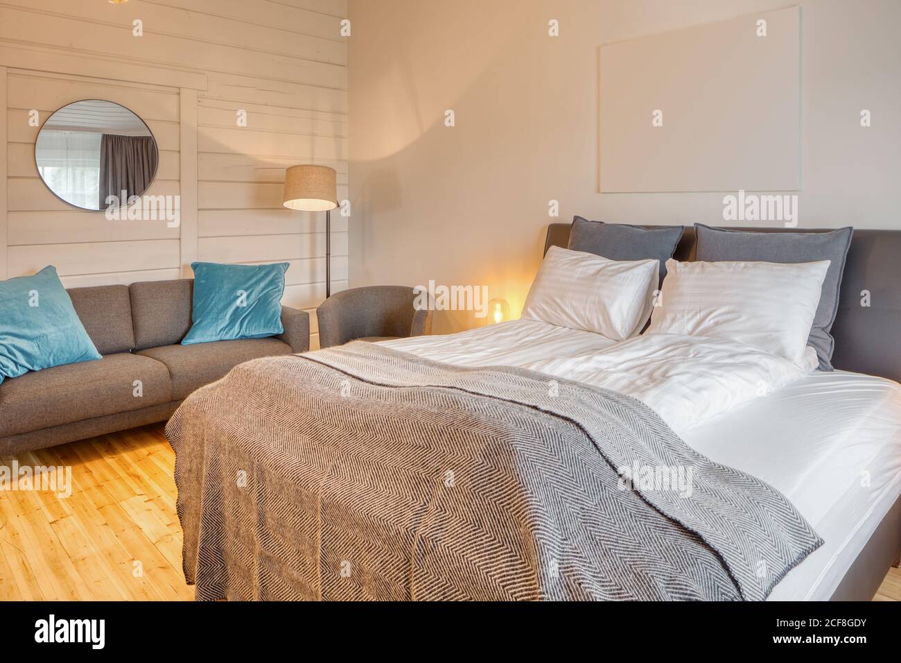 Chambre confortable avec murs en bois blanc et grand lit moelleux avec canapé gris confortable près de l'éclairage au sol Banque D'Images