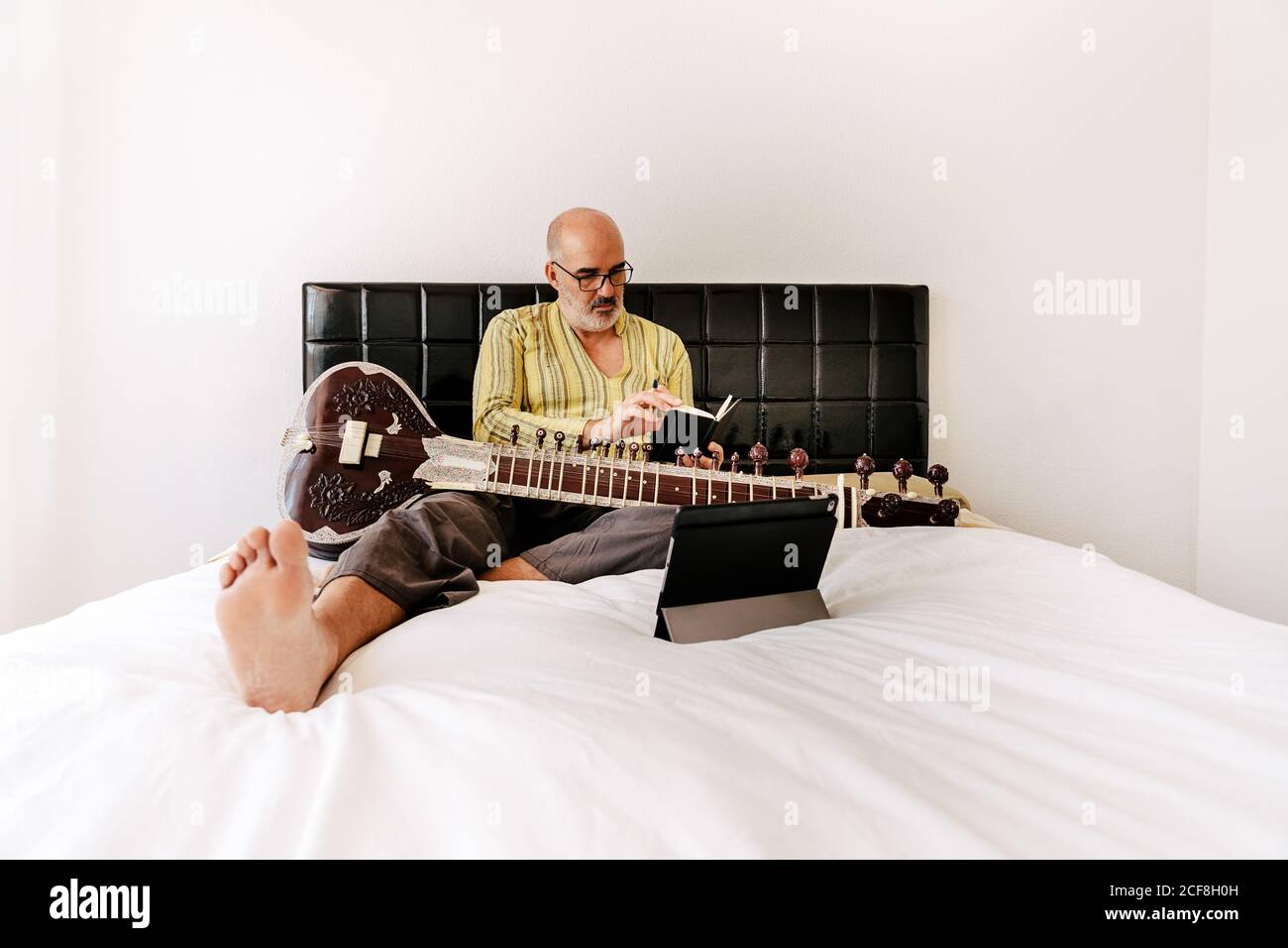 Homme âgé assis sur le lit avec un sitar et prenant des notes sur un ordinateur portable tout en regardant une leçon en ligne sur une tablette Banque D'Images