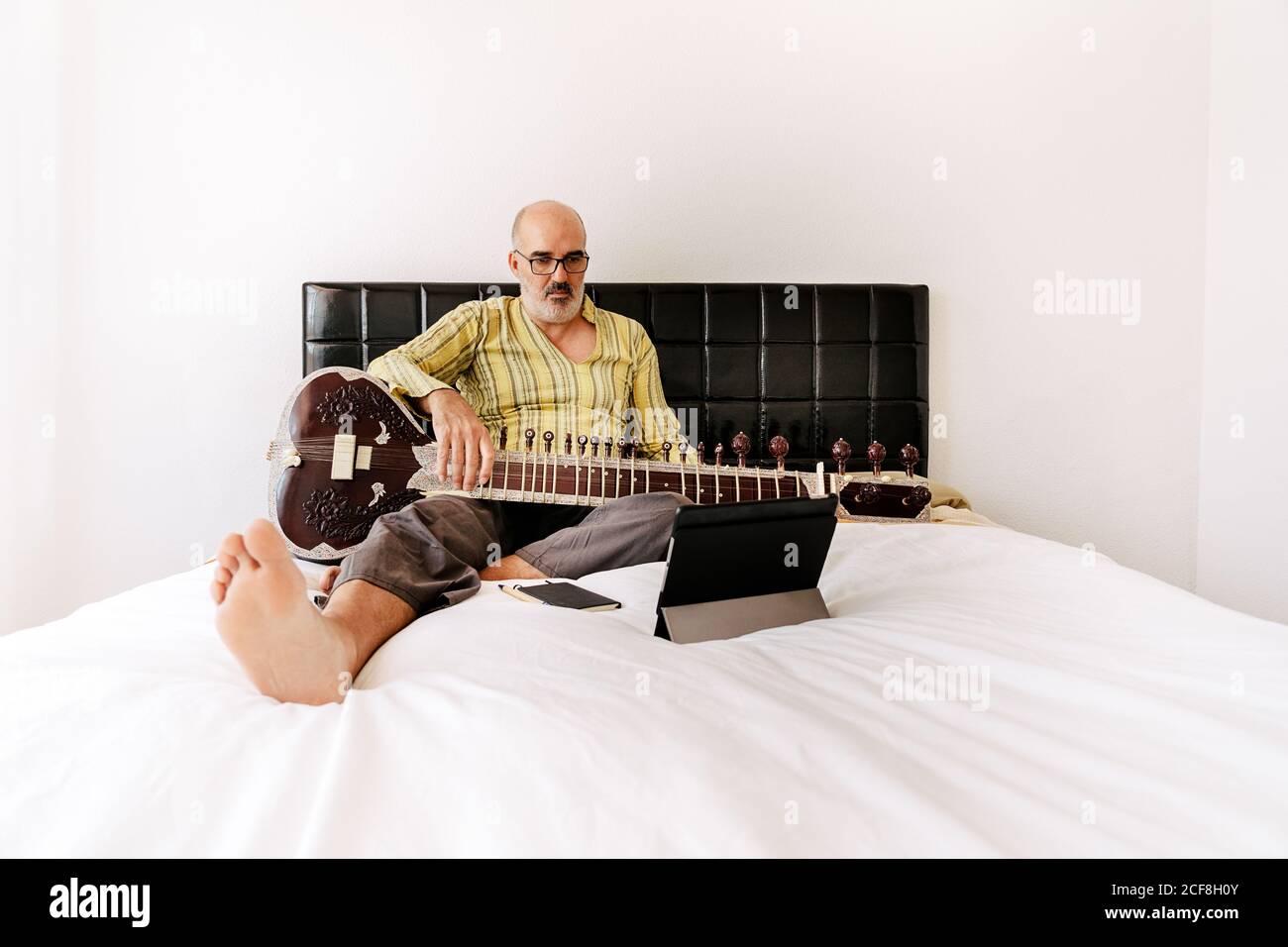 Homme âgé assis sur le lit avec un sitar et prenant des notes sur un ordinateur portable tout en regardant une leçon en ligne sur une tablette Banque D'Images