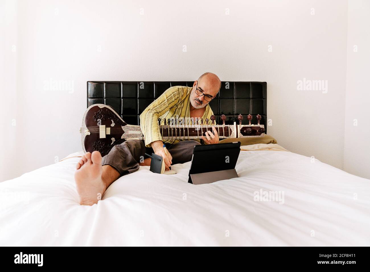 Homme âgé assis sur le lit avec un sitar et prenant des notes sur un ordinateur portable tout en regardant une leçon en ligne sur une tablette Banque D'Images