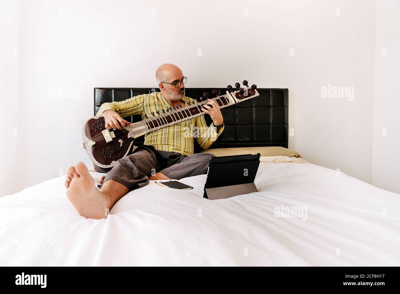 Homme âgé assis sur le lit avec un sitar et prenant des notes sur un ordinateur portable tout en regardant une leçon en ligne sur une tablette Banque D'Images