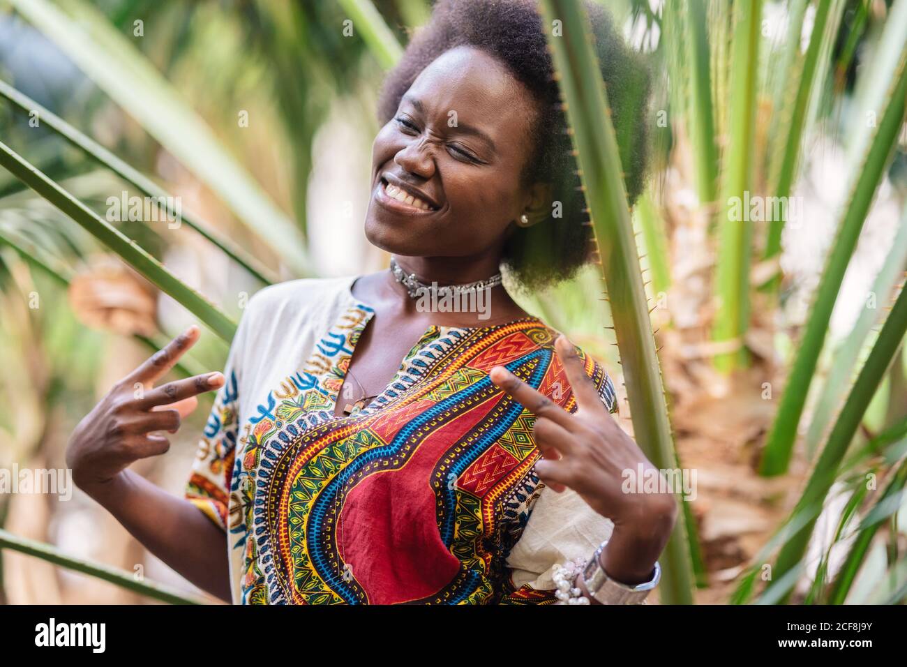 Bonne femme africaine aux cheveux bouclés souriant avec les yeux fermés et le signe V gestant tout en se tenant près des paumes le jour ensoleillé Banque D'Images