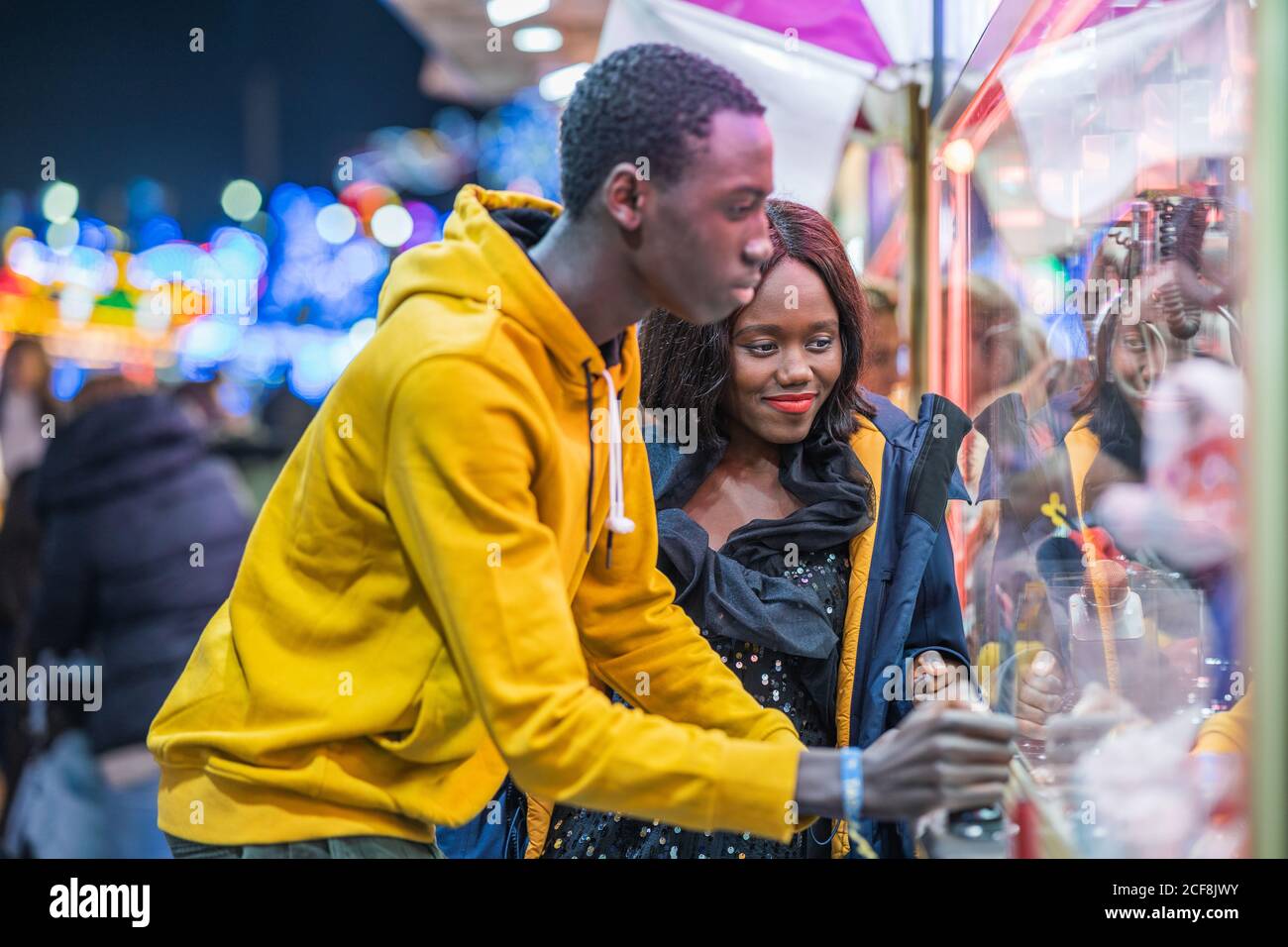 Femme afro-américaine souriant et regardant un petit ami attraper des jouets dans un jeu d'arcade tout en passant la soirée sur le funfair ensemble Banque D'Images