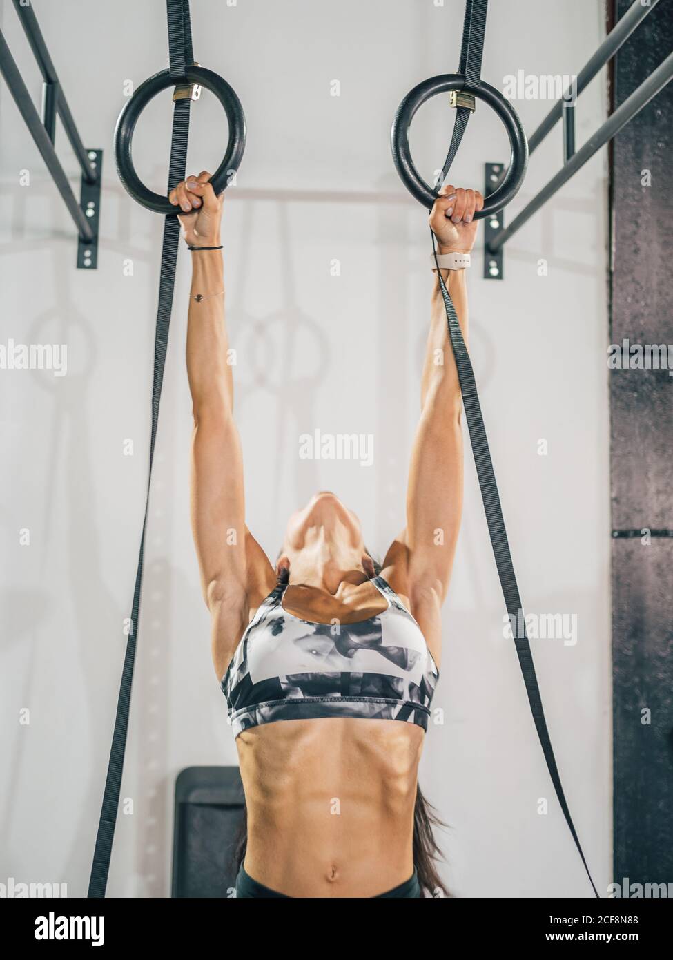 Faites de l'entraînement avec une athlète féminine adulte forte en vêtements de sport gris sur les anneaux de gymnastique noirs contre le mur blanc dans le club de sport Banque D'Images