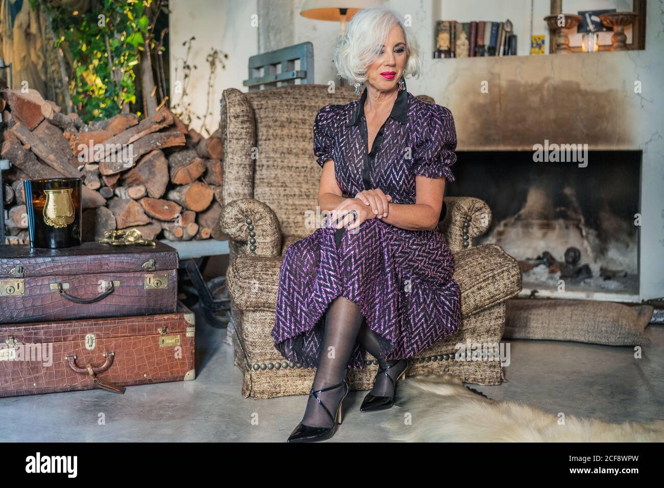 Calme et confiante dame dans l'élégante robe noire et pourpre et chaussures à talons hauts qui regardent et rêvent tout en étant assis fauteuil brun doux près des valises en cuir et de la vieille cheminée contre intérieur rustique vieilli Banque D'Images