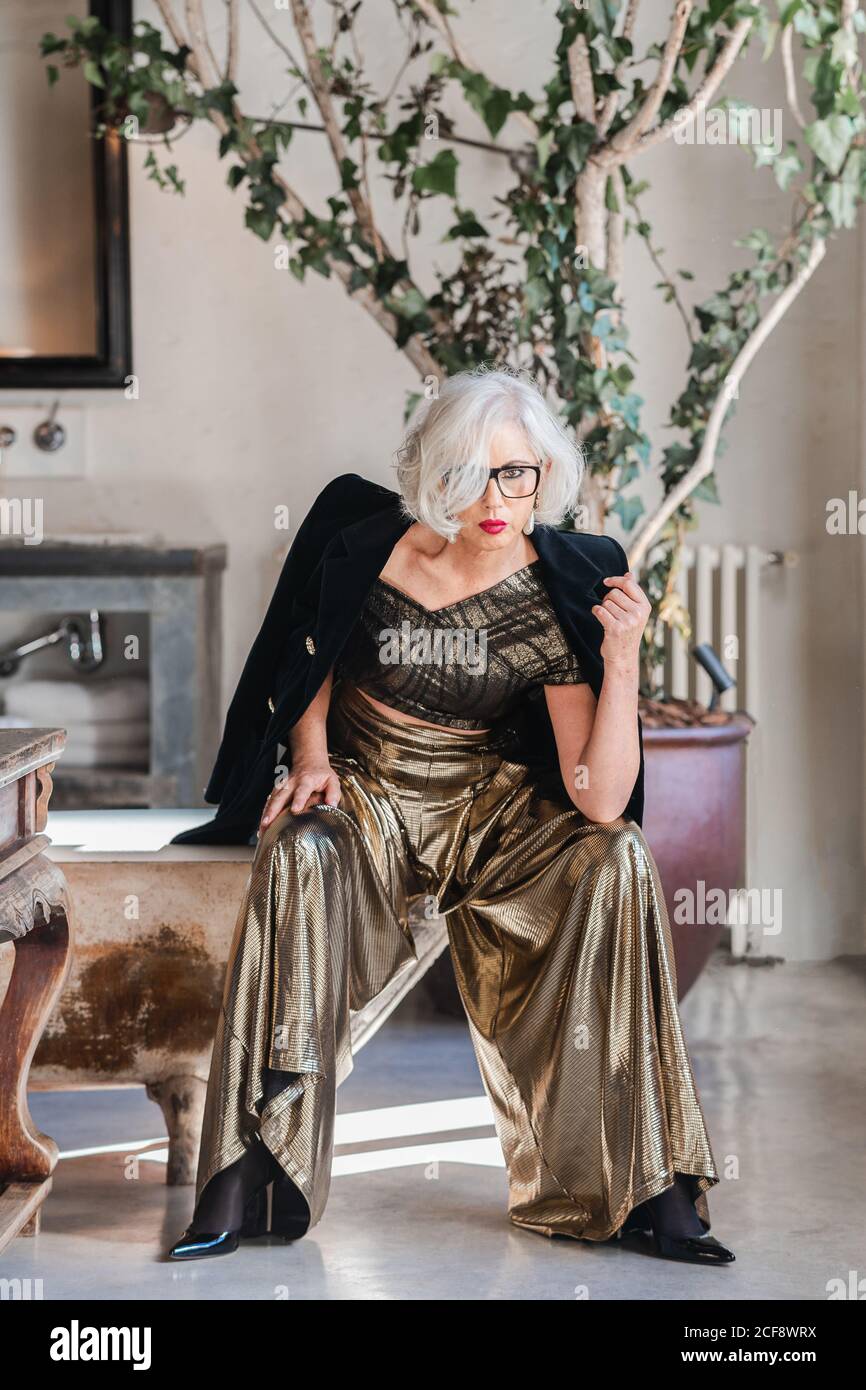 Calme senior confiante dame dans des lunettes et des vêtements élégants à la recherche à l'appareil photo tout en étant assis sur le bord de la baignoire de merde parmi les riches intérieur rétro avec meubles anciens et grand arbre à motif vert dans la chambre spacieuse de la maison de campagne Banque D'Images