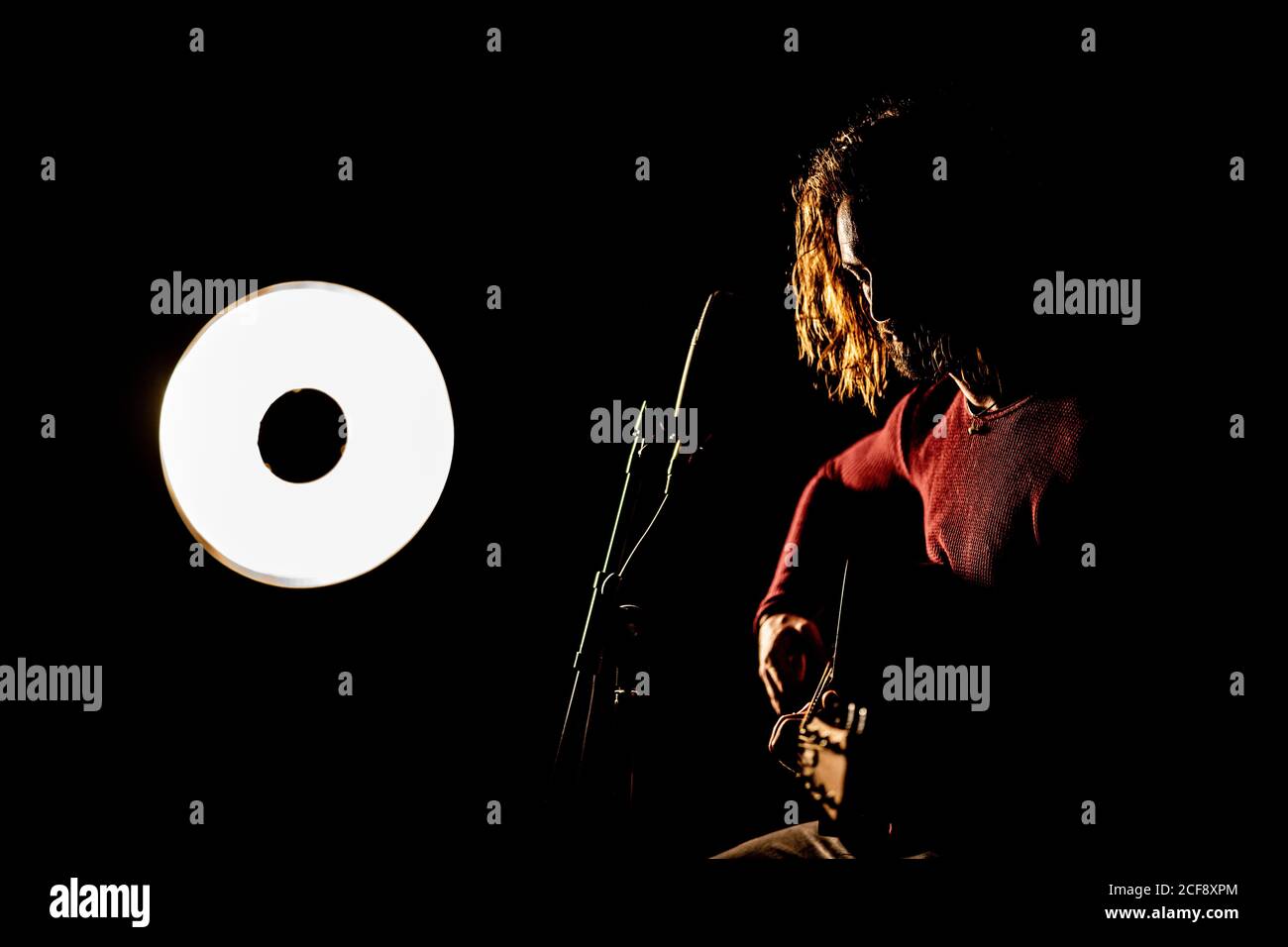 Guitariste mâle avec de longs cheveux assis dans un studio sombre à proximité microphone et lampe et guitare Banque D'Images
