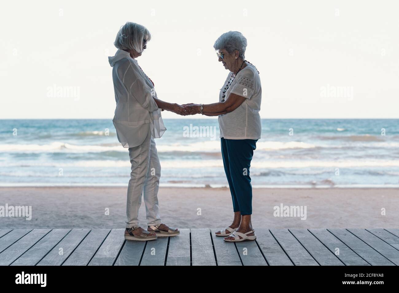 Vue latérale des femmes âgées aux cheveux blancs, calmes et gaies, qui soutiennent chacune d'elles autre et de tenir les mains sur la jetée de mer de la plage dedans heure d'été Banque D'Images