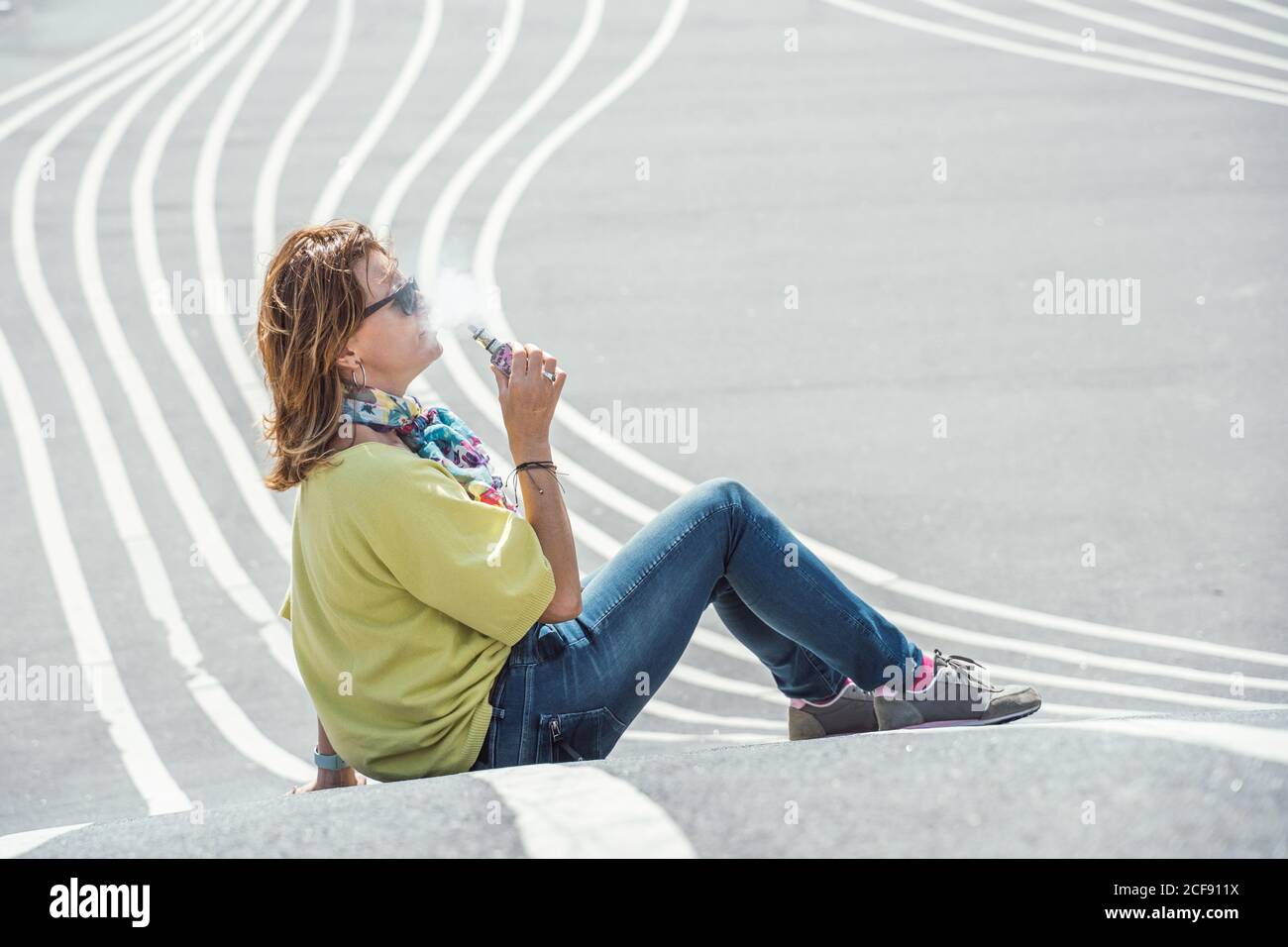 Vue latérale d'une femme qui fait froid chez un adulte dans des lunettes de soleil et un ensemble coloré qui exhalent de la vapeur tout en étant assise sur une route en asphalte tordant Banque D'Images