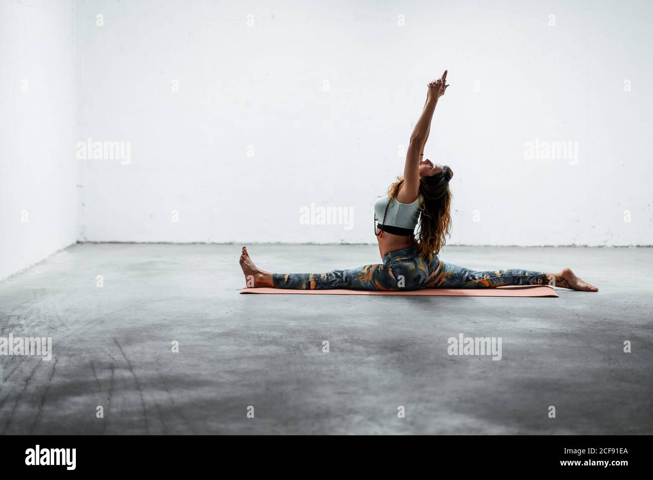 Vue latérale de la jeune femme flexible dans les vêtements de sport faisant devant séparer avec les bras relevés en étant assis sur le plancher Banque D'Images