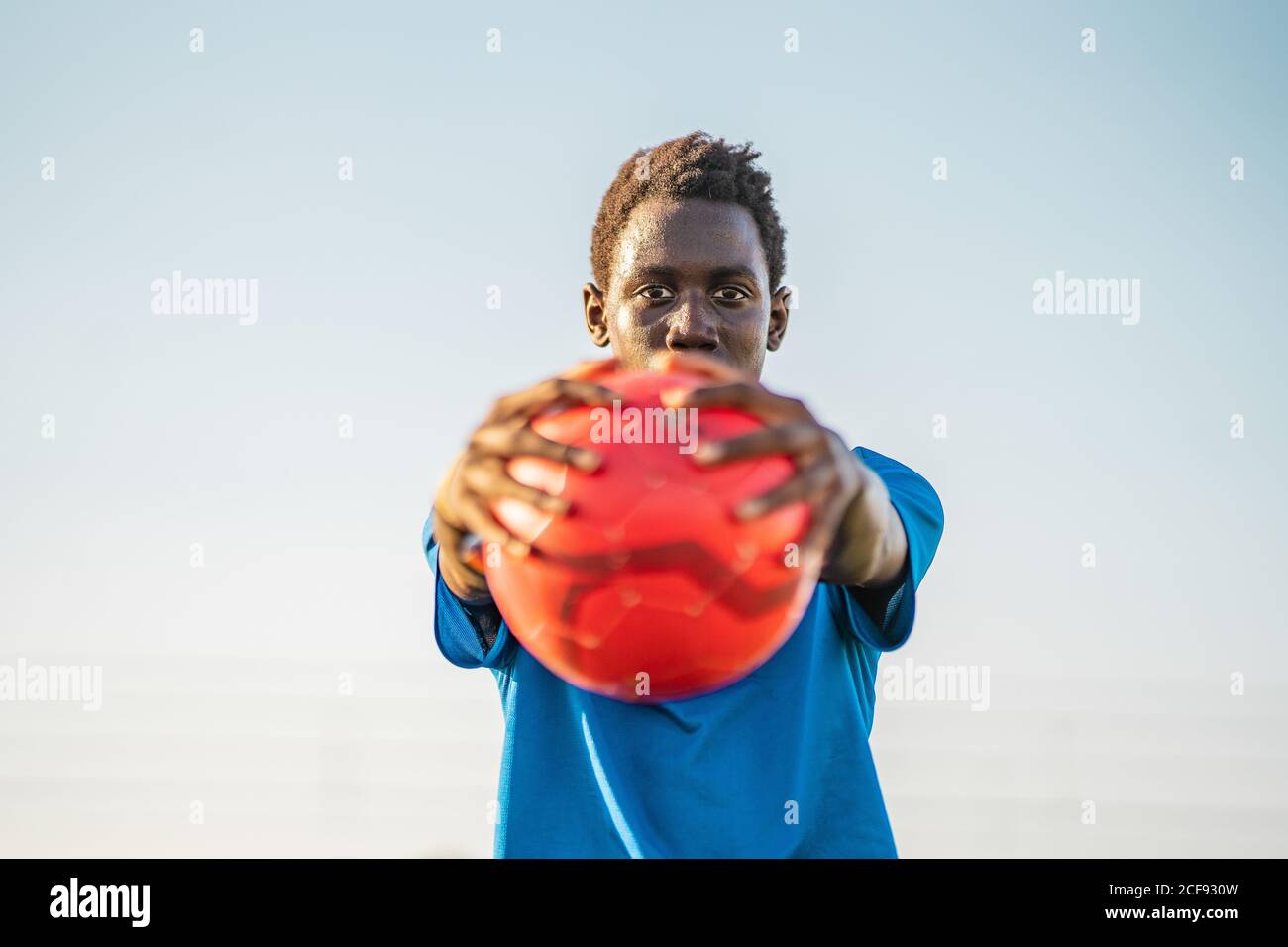 Adolescent noir en t-shirt bleu portant une balle rouge étirée bras et regarder la caméra contre un ciel sans nuages Banque D'Images