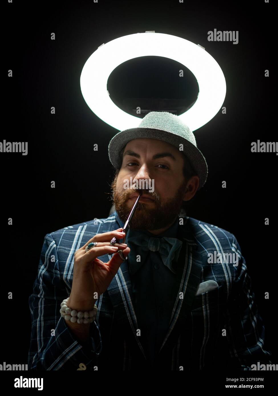 Homme barbu élégant avec des ciseaux planifiant soigneusement la coupe de cheveux sur le noir arrière-plan sous lumière blanche sur fond noir Banque D'Images