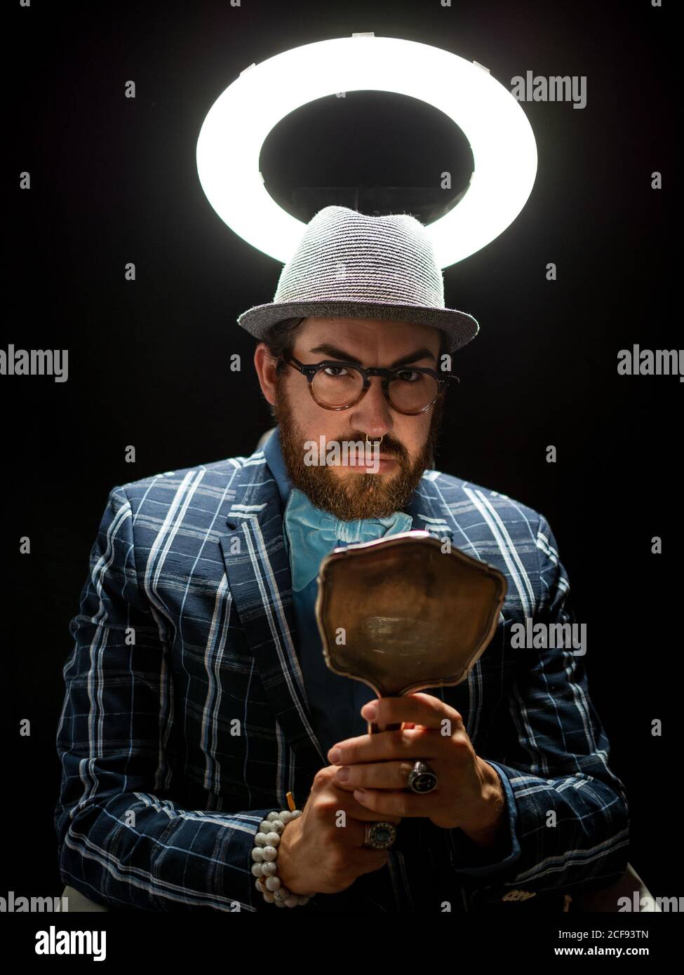 Homme barbu élégant dans des lunettes et un chapeau en admirant la réflexion à l'intérieur symétriser sous la lumière de cercle sur fond noir Banque D'Images