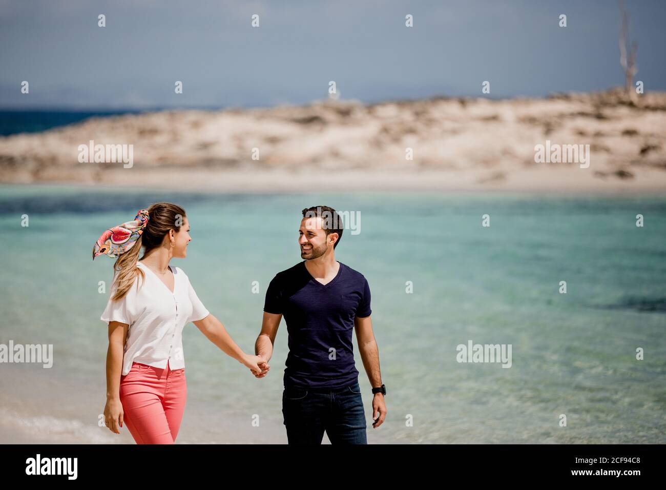 Homme souriant et femme marchant le long de la mer tout en tenant les mains et regardant l'un l'autre avec amour sur un arrière-plan flou Banque D'Images