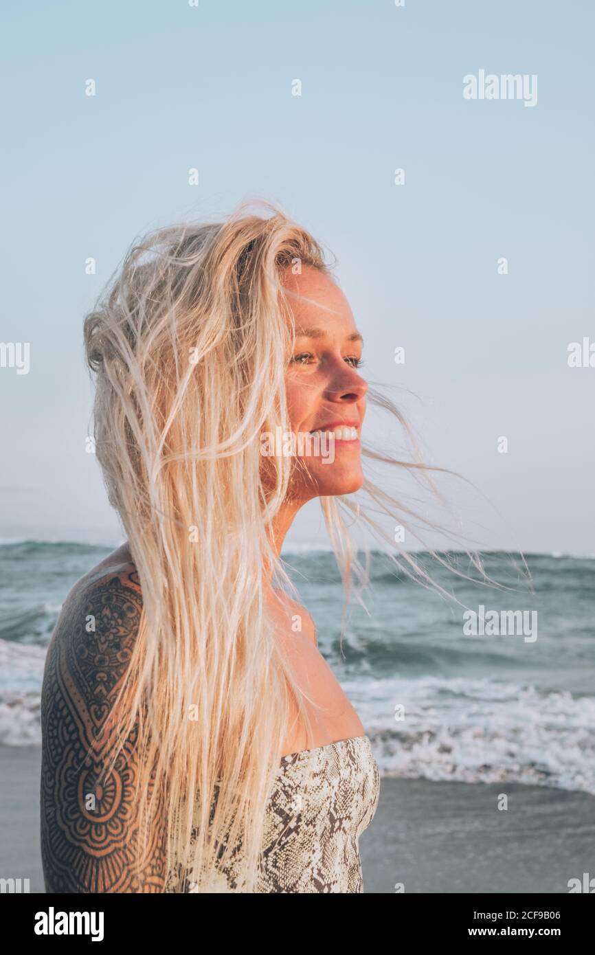Vue latérale d'une jeune blonde avec tatouage sur l'épaule souriant en vous tenant sur une plage de sable sur la mer et en regardant loin rêveur Banque D'Images