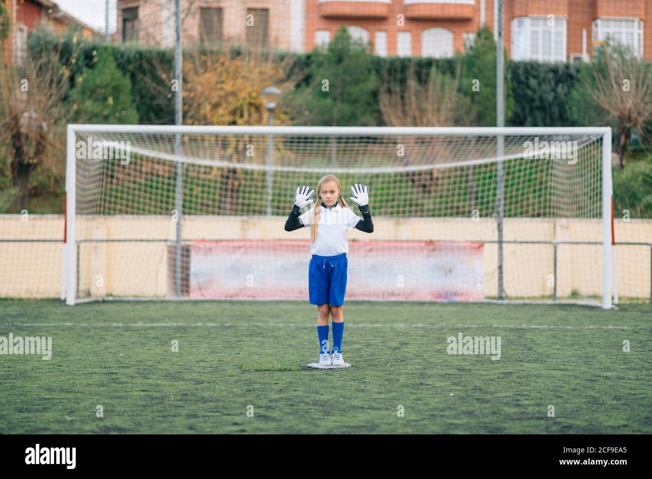 Gardien de fille de préadolescents sérieux en blanc et bleu uniforme défendant but de football en se tenant debout avec les bras levés seul sur le terrain pendant le match dans un stade moderne en plein air Banque D'Images