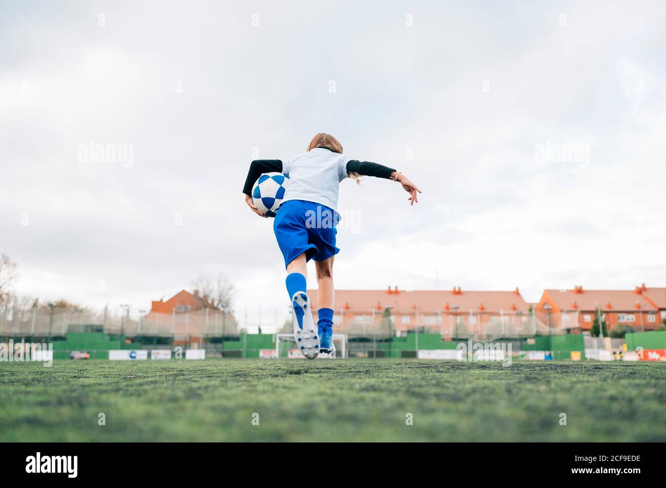 Vue arrière adolescente en uniforme blanc et bleu et chaussures de course à pied et préparation au ballon de football seul sur un terrain vert dans un club sportif contemporain Banque D'Images