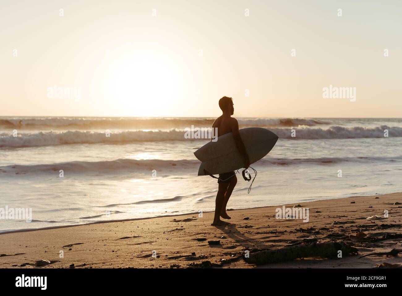 Vue arrière de la silhouette d'homme anonyme tenant une planche de surf pendant la marche le long de la plage de sable en été pendant le coucher du soleil Banque D'Images