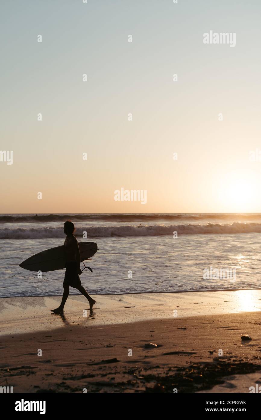 Vue latérale d'un homme anonyme tenant une planche de surf pendant la marche le long de la plage de sable en été pendant le coucher du soleil Banque D'Images