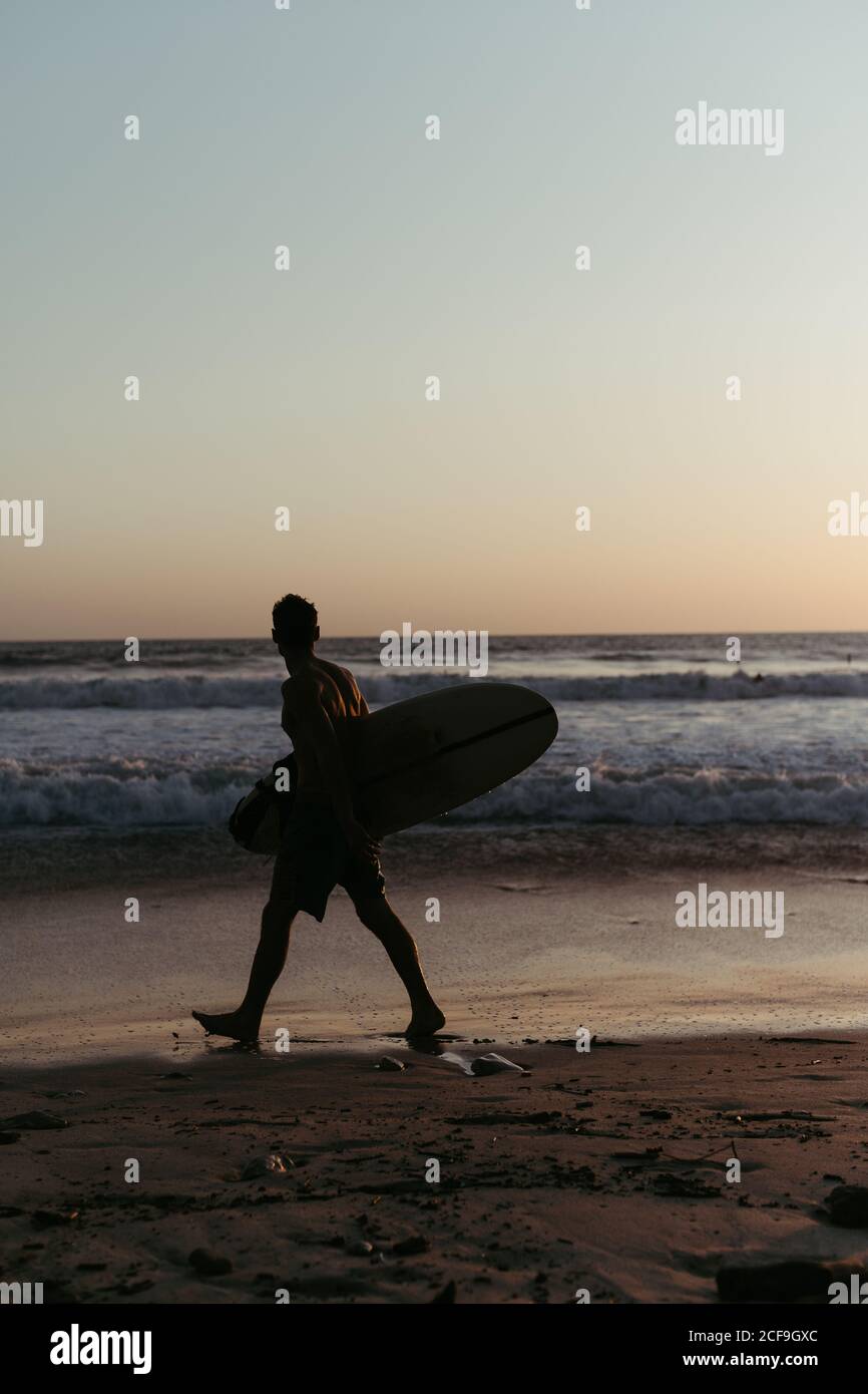 Vue latérale d'un homme anonyme tenant une planche de surf pendant la marche le long de la plage de sable en été pendant le coucher du soleil Banque D'Images