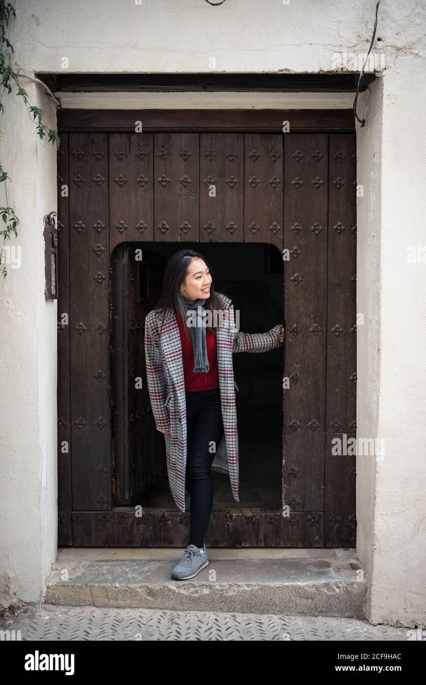 Une femme asiatique excitée dans un manteau décontracté souriant à la sortie de la petite porte et du vieux bâtiment et de regarder loin Albaicin à Grenade Banque D'Images