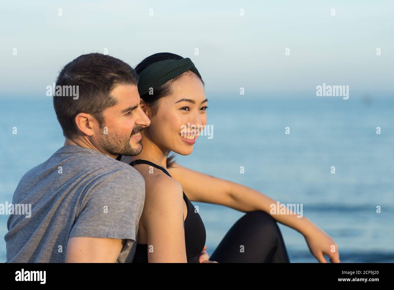 Vue latérale d'un jeune couple multiracial joyeux dans un salon sportswear sur une plage de sable, en vous reposant après un entraînement et en profitant du temps ensemble Banque D'Images