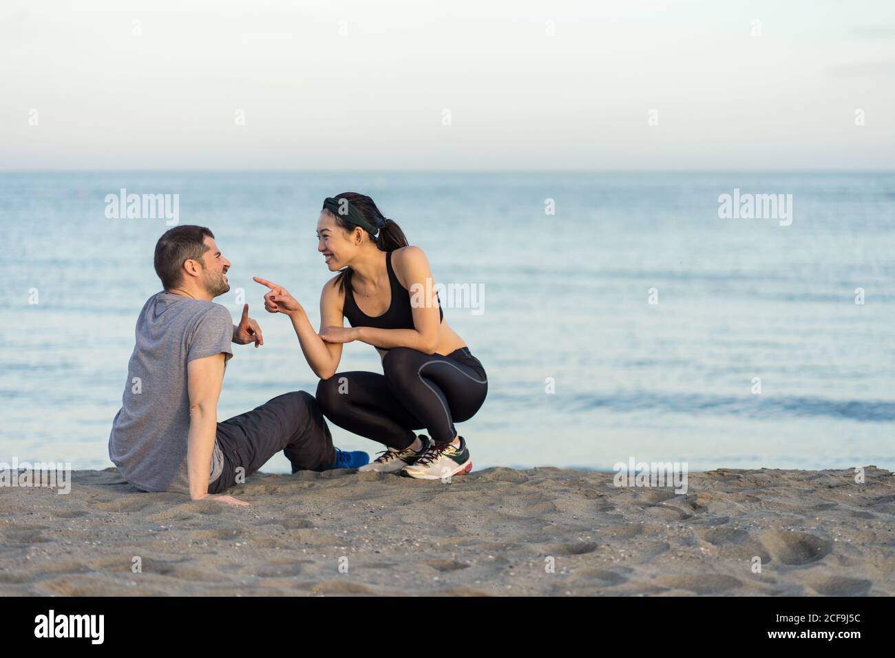 Vue latérale d'un jeune couple multiracial joyeux dans un salon sportswear sur une plage de sable, en vous reposant après un entraînement et en profitant du temps ensemble Banque D'Images