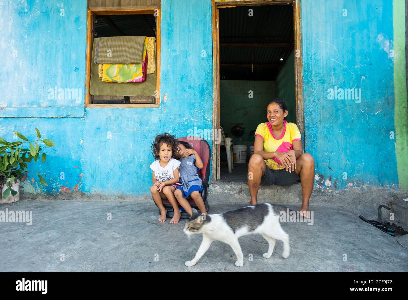 Dili, Timor oriental - 09 AOÛT 2018: Femme gaie et petites filles regardant la caméra tout en étant assis à la porte de la maison pauvre Banque D'Images