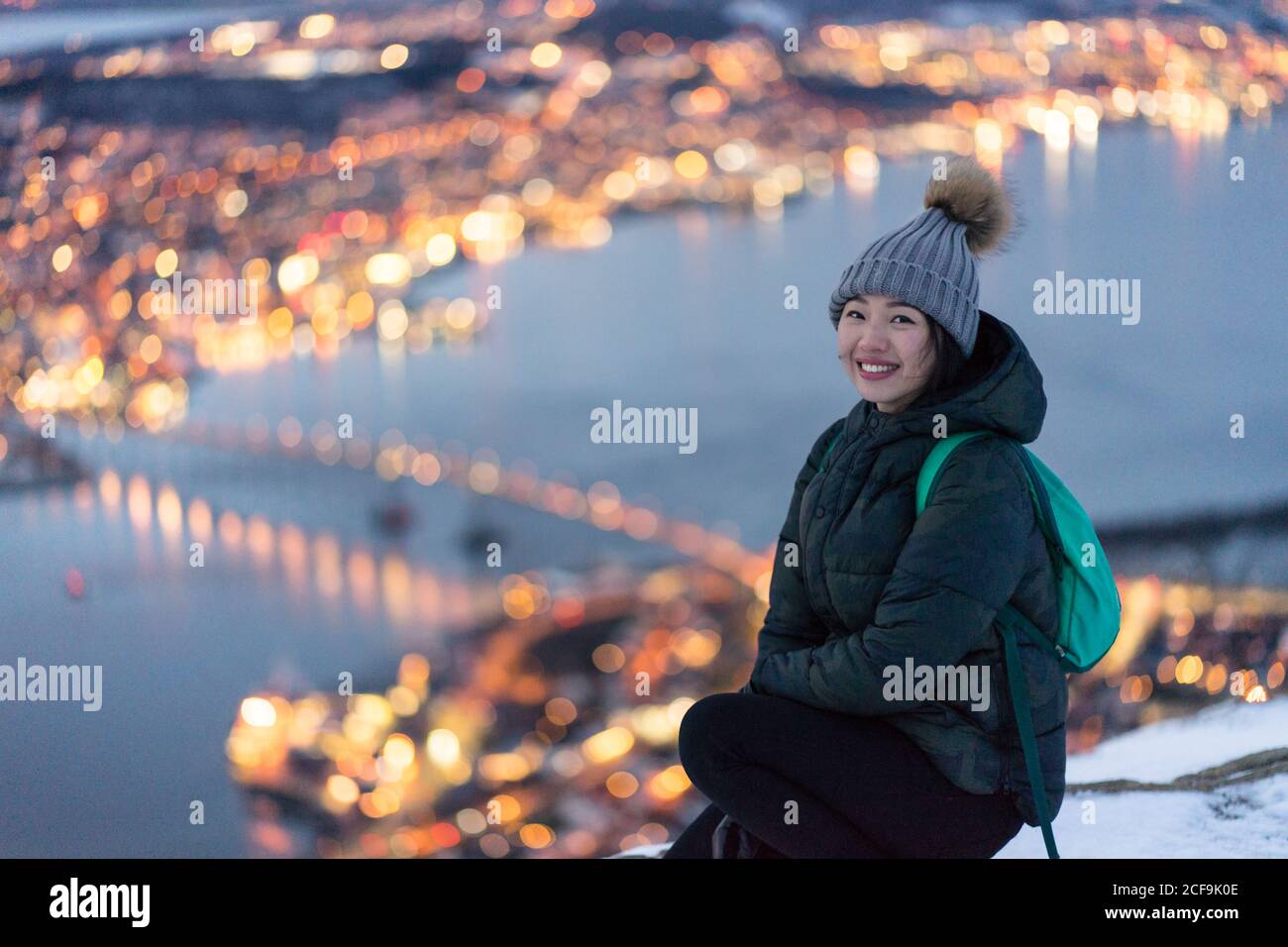 Jeune femme excitée en veste en duvet kaki et gris chaud chapeau regardant l'appareil photo et contemplant la vue d'hiver incroyable de ville située sur la côte en soirée Banque D'Images
