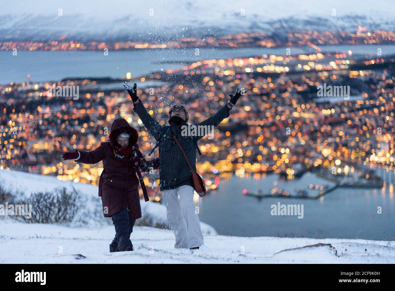 Un homme adulte heureux dans des vêtements chauds qui jettent de la neige comme un enfant et profiter de l'hiver tout en se reposant avec la femme ensemble à la périphérie debout sur une colline contre une vue incroyable des lumières dorées floues de la ville située sur l'île Banque D'Images