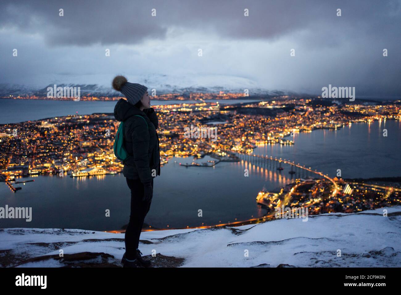 Jeune femme excitée en veste en duvet kaki et gris chaud chapeau regardant loin et contemplant la vue d'hiver incroyable de la ville situé sur la côte en soirée Banque D'Images